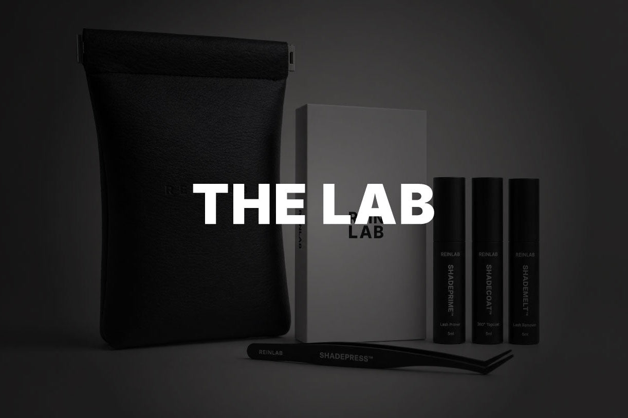 A dark studio product shot featuring a black pouch, a white box labeled 'THE LAB', three slim black tubes, and a black applicator wand arranged on a dark surface against a dark gray background.